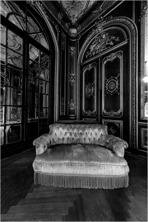 A vintage tufted sofa with fringe in an ornate room with dark walls and large windows.