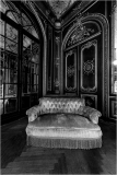 A vintage tufted sofa with fringe in an ornate room with dark walls and large windows.