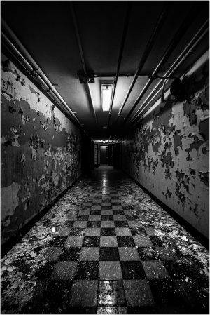 Long corridor with peeling walls and checkered floor, illuminated by overhead lights.
