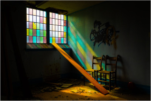 Abandoned room with colorful stained glass windows and a wooden plank leaning against them.
