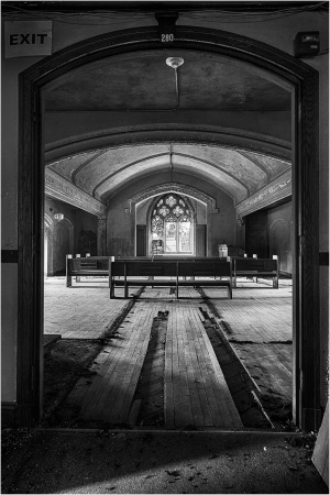 A dimly lit room with worn wooden floors, an archway leading to empty benches.