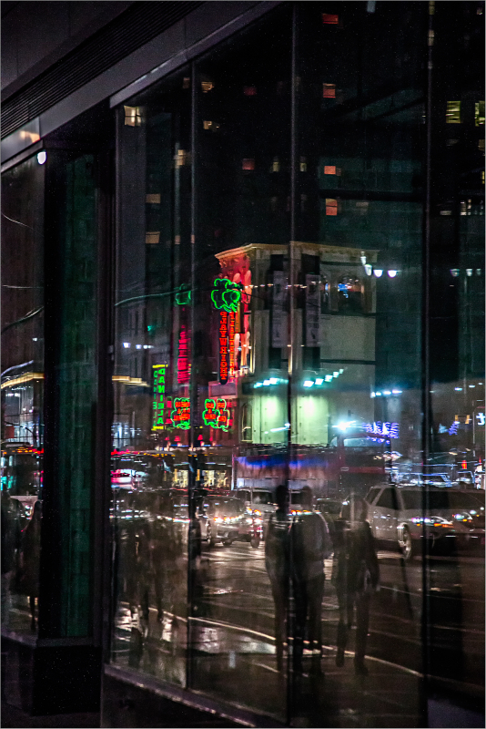 City lights reflected in a glass building at night, with people walking by.