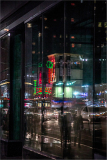City lights reflected in a glass building at night, with people walking by.