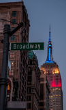 Broadway street sign with the Empire State Building lit in red, white, and blue in the background.