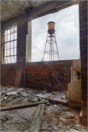 A weathered interior with debris, framing a distant water tower against a cloudy sky.