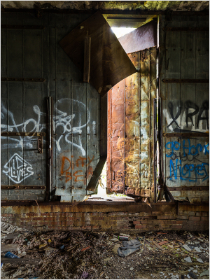 A weathered door stands ajar, revealing light and shadows in a graffiti-covered space.
