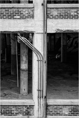 Black and white image of industrial pipes along a concrete pillar in a dilapidated building.