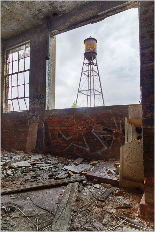 A weathered interior with debris, framing a distant water tower against a cloudy sky.