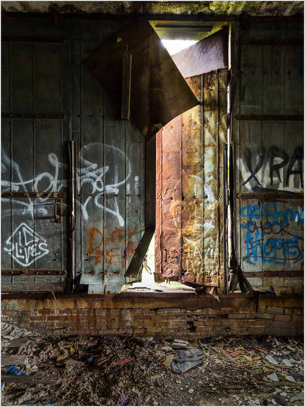 A weathered door stands ajar, revealing light and shadows in a graffiti-covered space.
