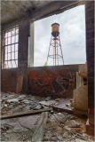 A weathered interior with debris, framing a distant water tower against a cloudy sky.