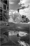 A desolate urban landscape with a puddle reflecting a watchtower and abandoned buildings.