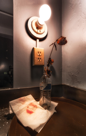 A faded rose in a bottle beside a water bottle and crumpled napkin under a dim light.