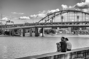 Two people sit by a river with a bridge in the background on a sunny day.
