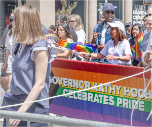 A parade scene with people celebrating Pride, featuring a banner for Governor Kathy Hochul.