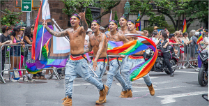 Four shirtless men cheerfully wave rainbow flags during a parade celebration.