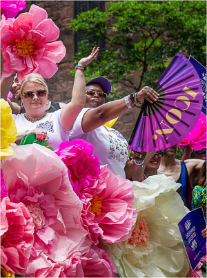 A lively Pride parade scene with people joyfully waving fans and surrounded by oversized flower decorations.