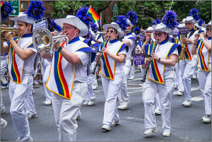 Marching band in white uniforms, colorful sashes, playing instruments at a Pride parade.