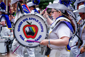 A marching band member holds a drum with a colorful logo and vibrant attire during a parade.