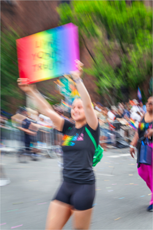 Person holds a rainbow sign at a lively parade, surrounded by cheering crowds.