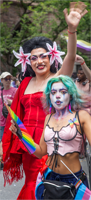Two performers in vibrant outfits wave at a parade, one in a red dress and the other in a pink top with rainbow accessor