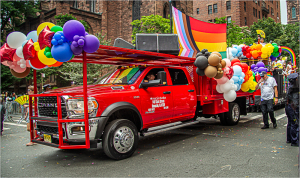 A red truck adorned with colorful balloons and a rainbow flag at a parade.