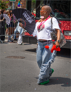 A person in a white top and fringed jeans holds a fan, walking at a festive parade.