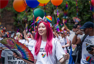 A vibrant parade scene with a person in a rainbow-themed outfit and pink hair, holding a fan.
