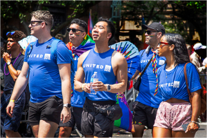 Group of people in blue shirts celebrating at a parade with colorful fans and rainbow accessories.