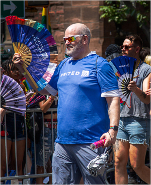 Man in blue shirt holds colorful fans at a parade with crowd behind him.