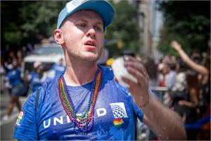 Person wearing a blue cap and beads at a vibrant parade, with water splashing.