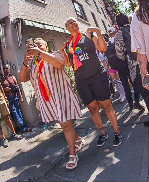 Two joyful women celebrate Pride Day in NYC, adorned with vibrant rainbow sashes.