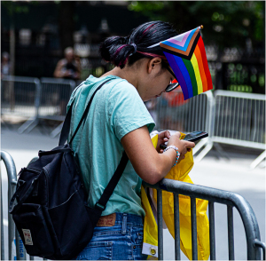 Individual at PRIDE NYC, wearing a rainbow flag hat, focused on a smartphone.