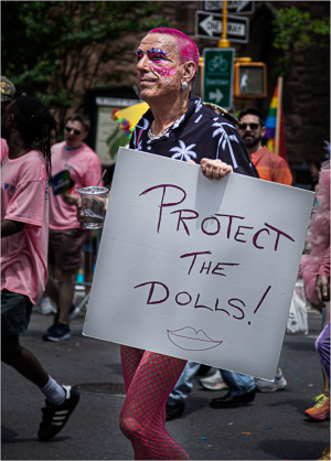 Individual with pink hair holds a sign saying 'Protect the Dolls!' at a vibrant parade.