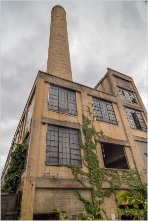 An abandoned industrial building with a tall smokestack and ivy creeping up its walls.