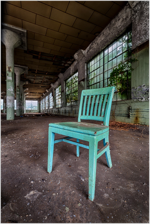 A quirky turquoise chair stands alone in a spacious, worn-out industrial room.