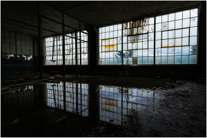 Abandoned interior with large, dirty windows reflecting on a dark puddle.