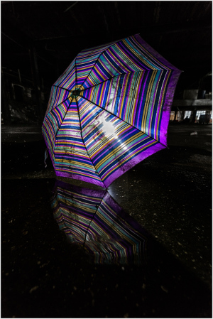 A vibrantly striped umbrella stands alone, casting its eerie reflection on darkened ground.