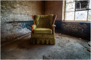 An abandoned green armchair sits alone in a dilapidated space, shadows dance around it.