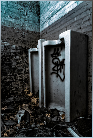 Two gritty urinals stand in a shadowy, crumbling room, graffiti marking their decay.