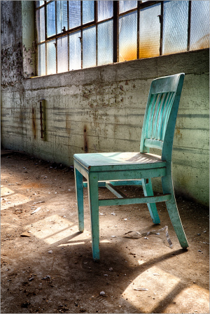 A lonely teal chair stands against a wall with large windows, light streaming in, dust dancing around.