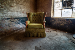 An abandoned green armchair sits alone in a dilapidated space, shadows dance around it.