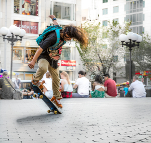 A skateboarder with wild hair performs a trick in a bustling city plaza.