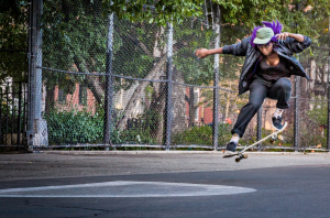 A skateboarder performing a jump with purple hair and a green hat.