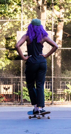 A person with vibrant purple hair skateboards, hands on hips, exuding confidence.