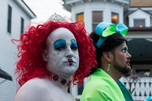 Two performers in vibrant costumes, one with red curls and dramatic makeup, the other in green attire.