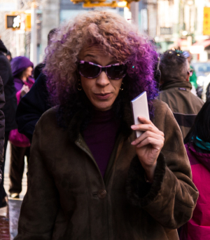 A person with curly purple hair and sunglasses holds a card, standing in a bustling street.