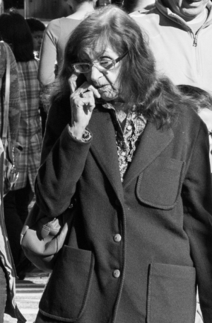 An elderly woman in a coat talks on her phone while walking through a crowd.