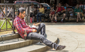 A man in a red plaid shirt sits on steps, reading a book in a lively park.