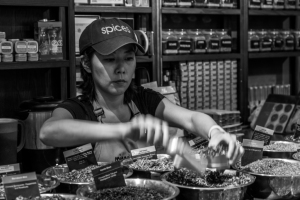 A focused woman in a cap measures spices in a bustling shop, surrounded by jars and bowls.