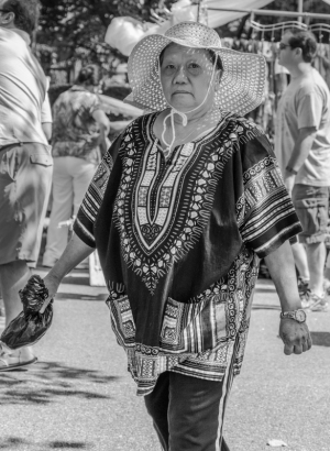 A woman in a patterned dress and wide-brimmed hat walks through a crowd.
